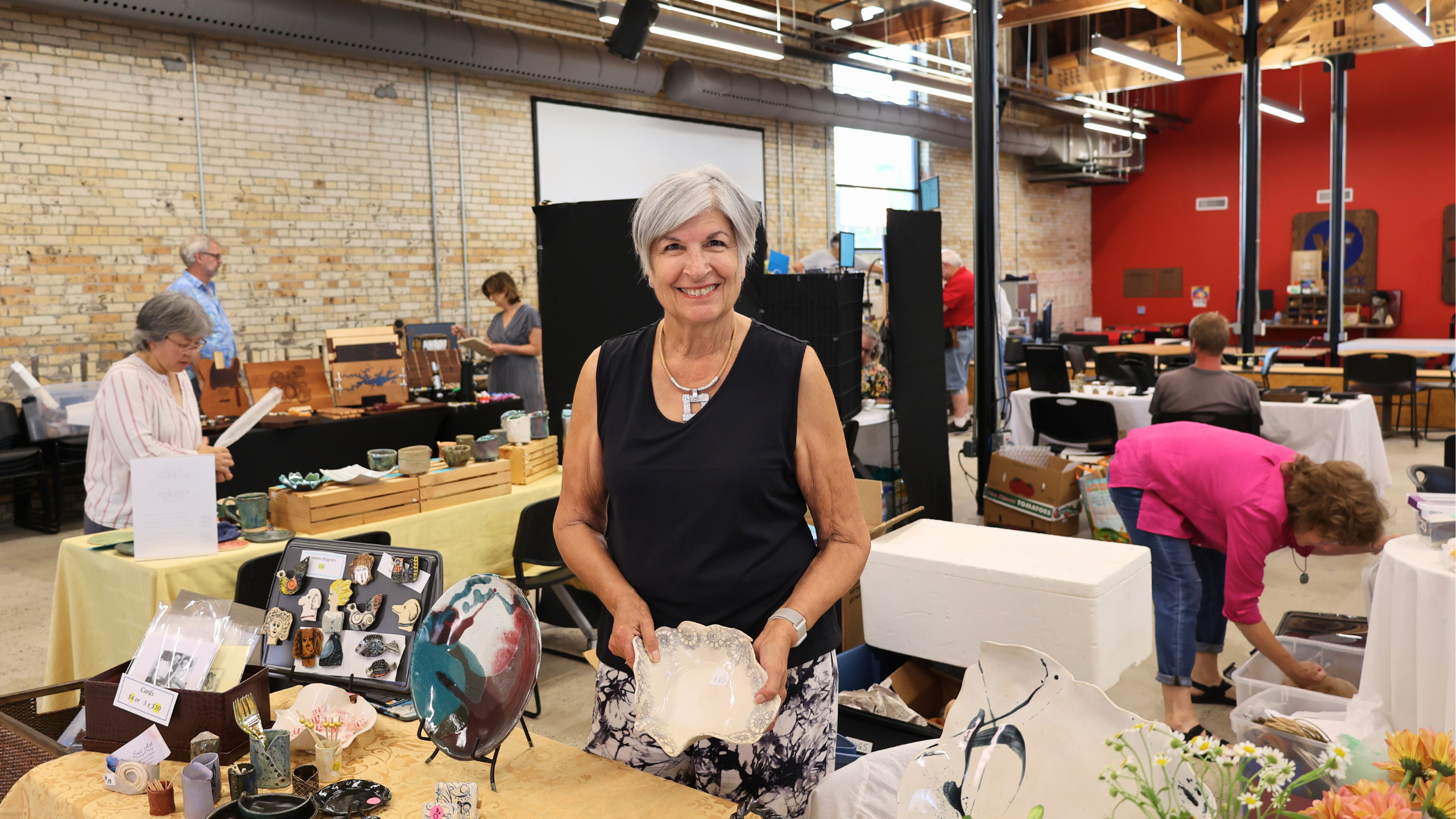 Susan Peterson setting up her booth for 3rd Friday Artwalk, holds a ceramic piece at an art fair with various artworks and people in the background.