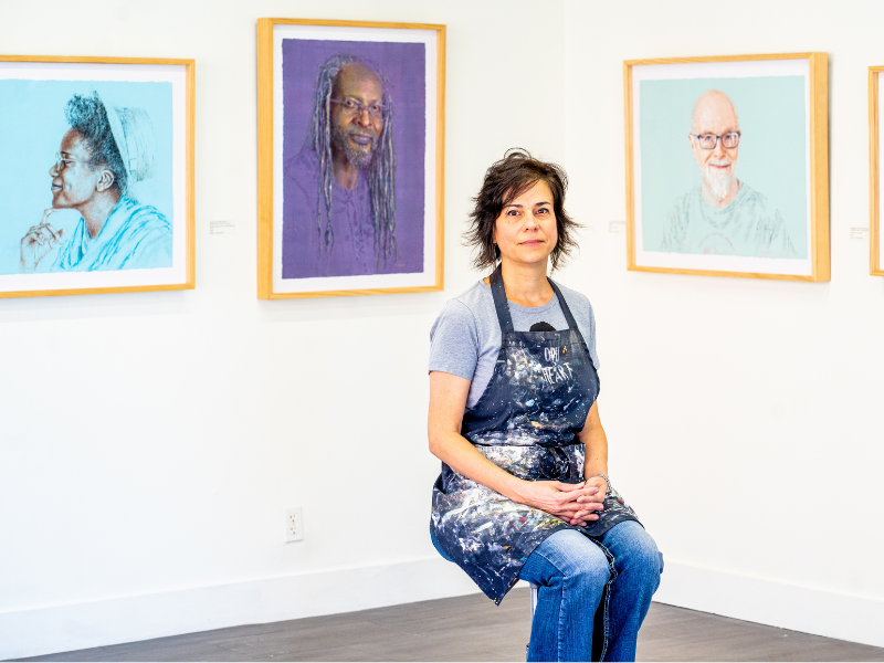 Virmarie DePoyster poses in front of her pieces wearing her colorful artist's apron.