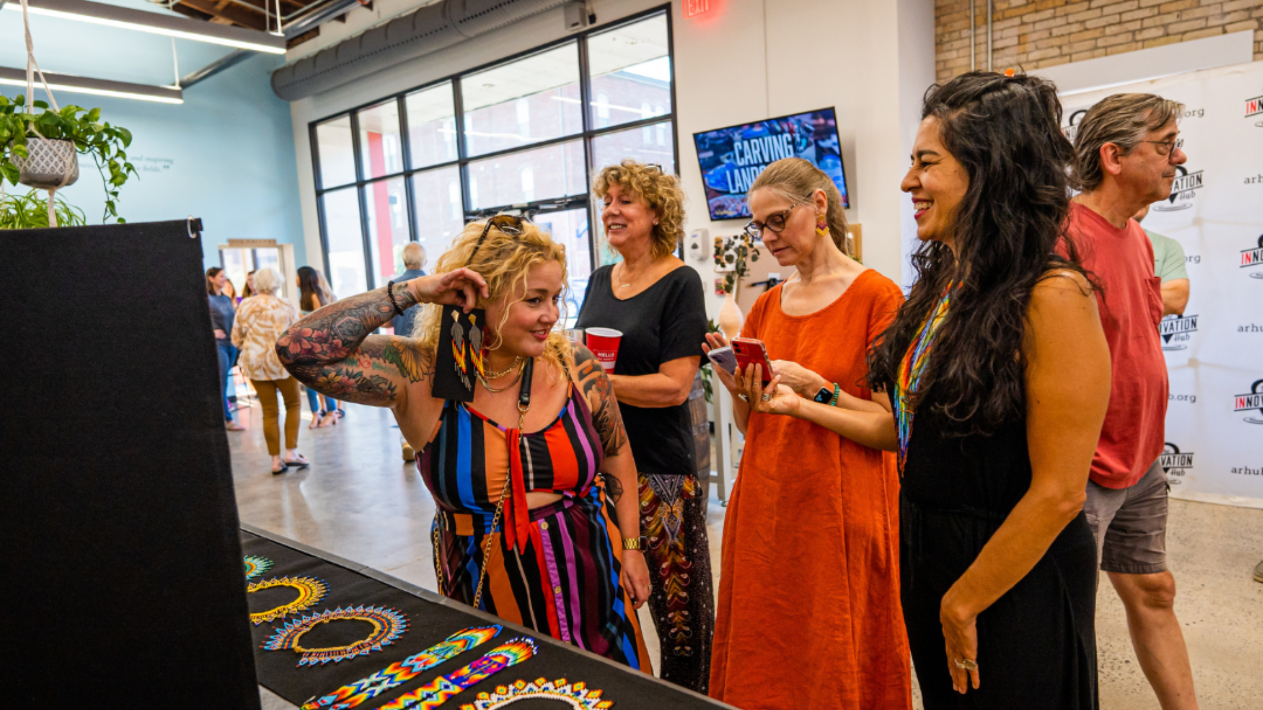 A group of women, dressed in vibrant clothing, admire the hand beaded and vibrant jewelry at Maria's booth inside the Innovation Hub.  One woman holds a card up to her ear with beaded earrings, modeling the look in front of a mirror.
