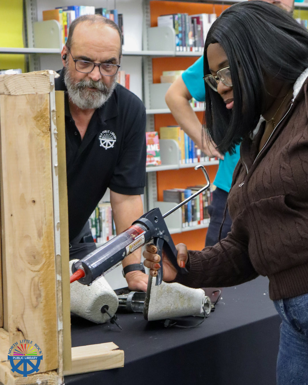 Two people working on a project involving a wooden box, caulk, and tools in a library setting.