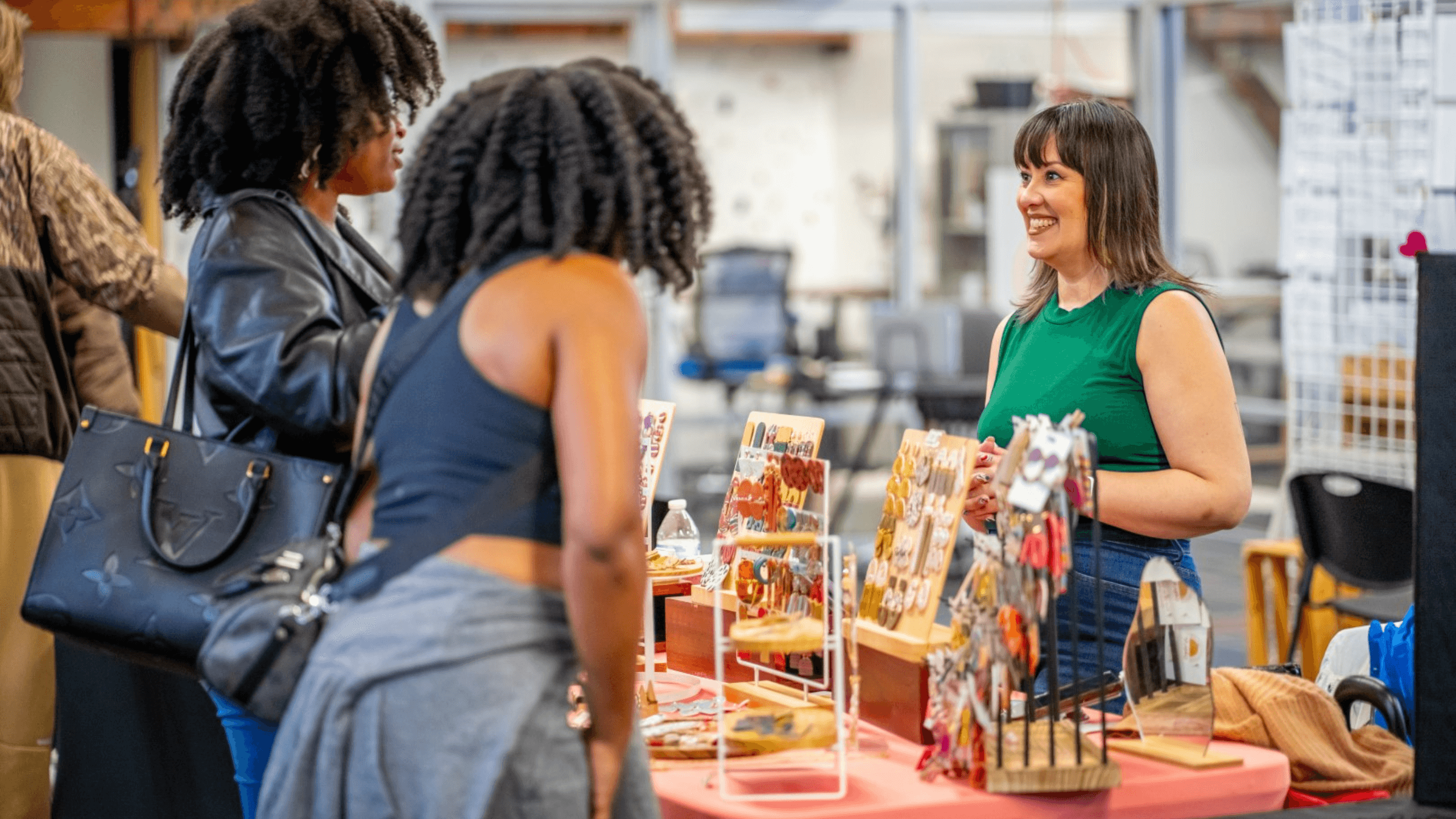 Two women are interacting with Dani Golden at her booth inside the Hub during the monthly indoor craft fair, with various items on display.