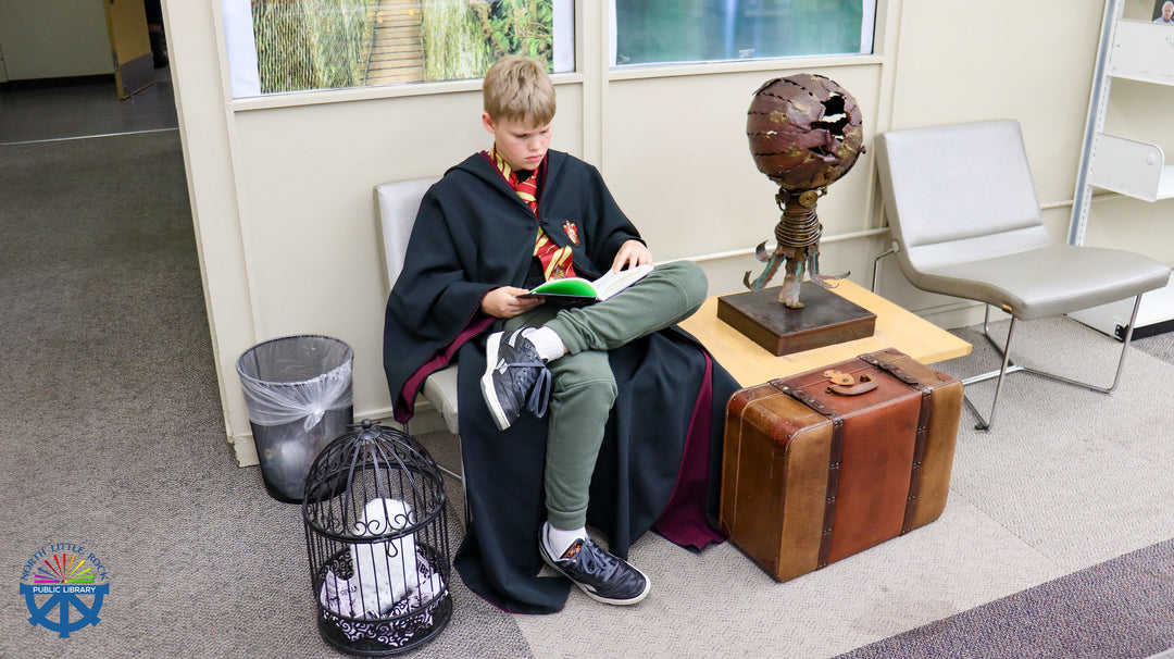 A child wearing a howarts wizard robe reading a book while next to him sits a suitcase and on the other side a birdcage with a white owl - obiously on the way to hogwarts.