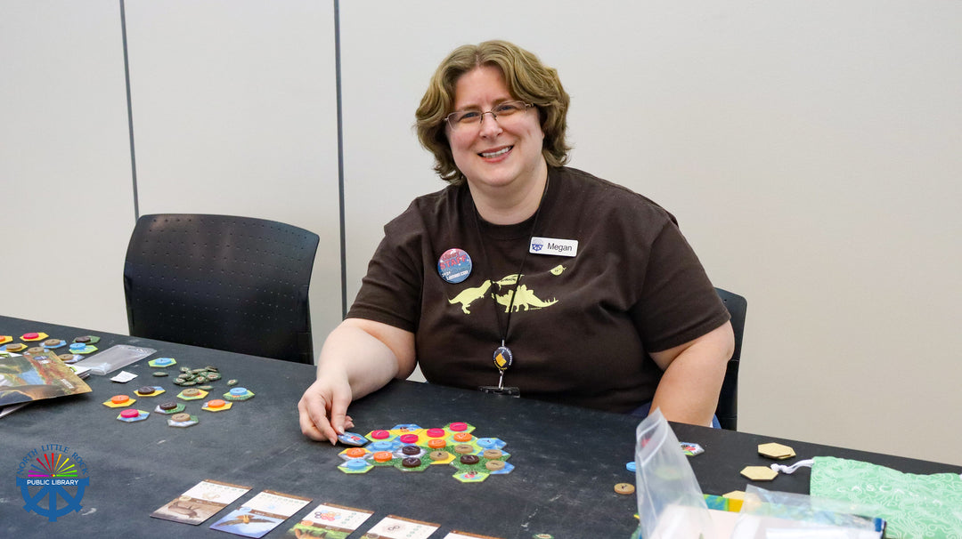 Library staff, Megan, sitting at a table with board game components, wearing a brown t-shirt with a dinosaur logo.