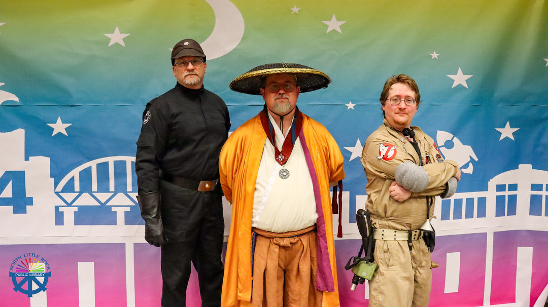 Three people in costumes standing in front of a colorful Laman Con 2024 banner with a cityscape design. First, second, and third prize costume winners.